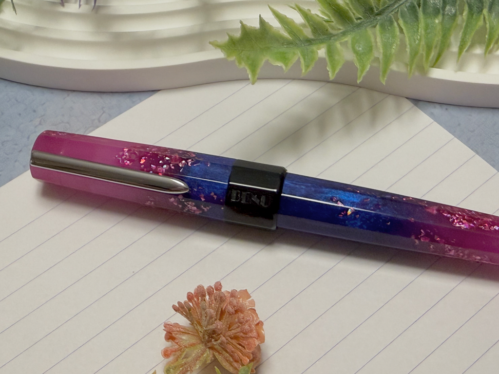 A resin pen with a pink-to-blue gradient and embedded glitter, resting on lined paper with a small dried flower and green leaves in the background.