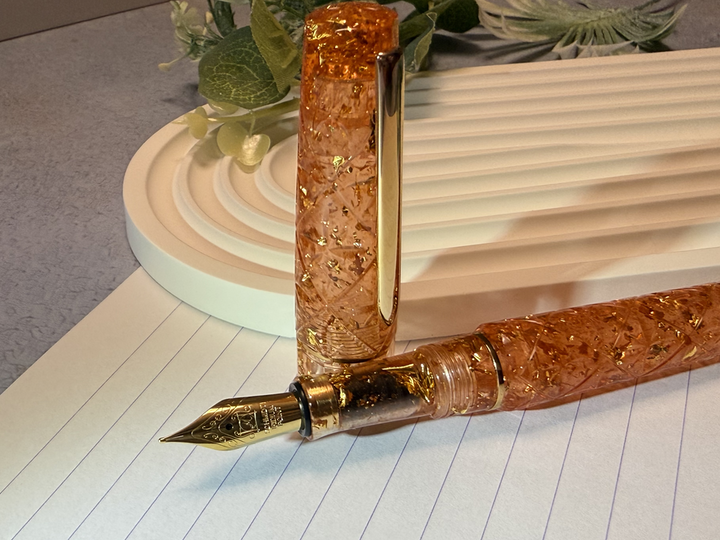 An amber-orange translucent fountain pen with gold-toned metal accents and embedded glitter, shown with its matching cap standing upright on lined paper.