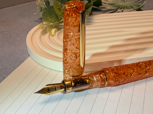 An amber-orange translucent fountain pen with gold-toned metal accents and embedded glitter, shown with its matching cap standing upright on lined paper.